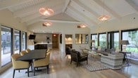 This single-story space showcases an open-plan layout that seamlessly connects multiple functional zones, creating a spacious and cohesive environment. The most striking architectural feature is the vaulted ceiling with exposed white-painted beams, which adds significant height and a sense of airiness throughout the main area. Three rose-tinted pendant lights hang from the ceiling, casting a warm, ambient glow that complements the natural light streaming in through large black-framed windows overlooking lush greenery outdoors.

The flooring throughout is light-toned hardwood, contributing to the bright and welcoming atmosphere. The lounge area is furnished with a mix of accent chairs and a plush sofa arranged around a glass coffee table, anchored by a patterned area rug. Adjacent to the lounge, a dining area features a dark table paired with upholstered chairs. Toward the rear, a kitchenette is equipped with dark cabinetry, counter seating, and modern appliances. Recessed alcoves along one wall provide additional intimate seating nooks, enhancing the space's versatility and functionality.