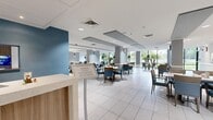 Located at 1099 N McMullen Booth Rd in Clearwater, Florida, this expansive single-floor dining facility offers a generously proportioned, open-concept layout designed for comfort and functionality. The space is characterized by its clean, contemporary aesthetic, featuring large-format white tile flooring, white ceilings with recessed lighting, and a refined color palette of soft blues, grays, and warm wood tones. Structural columns are integrated seamlessly into the design, accented with muted blue-gray paint that complements the upholstered seating throughout. A defining feature of the space is its extensive floor-to-ceiling glass window walls, which flood the interior with natural light and provide sweeping views of the surrounding lush, green landscape. Dining tables of varying configurations—round and rectangular—accommodate both intimate and group settings, with coordinating blue cushioned chairs lending a cohesive, welcoming feel. A private dining alcove offers a more secluded setting, complete with curated wall art and acoustic ceiling panels. Wide, well-lit corridors connect the main dining area to additional seating zones and service areas, including a host stand near the entrance. Framed botanical artwork decorates the walls throughout, enhancing the polished, upscale ambiance of this thoughtfully designed communal dining destination.