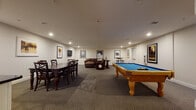 Located at 2-48 Grove Ave in Cedar Grove, NJ, this single-floor community recreation room is thoughtfully designed for socializing and leisure. The space is anchored by a full-size billiards table with a vibrant blue felt surface and solid wood legs, positioned at the center of the main open area, offering ample room for gameplay. Recessed lighting throughout provides consistent, comfortable illumination across the patterned carpet flooring.

A dedicated lounge area features plush leather sofas and a recliner, arranged for relaxed conversation or casual gathering. The walls are adorned with a curated collection of framed classic movie posters — including iconic titles such as Jaws, Reservoir Dogs, Rocky, and The Professional — lending the space a cinematic personality.

A formal dining area accommodates a dark wood table with matching cross-back chairs, ideal for group meals or meetings. Adjacent to the dining space is a kitchenette equipped with white cabinetry, a dark stone countertop, and a sink, providing convenient access to refreshments. A bistro-style high-top table and stools offer an additional casual seating option. The overall ambiance is polished yet inviting, making this a versatile amenity space well-suited for entertaining and community gatherings.