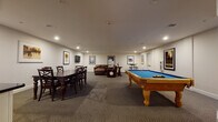 Located at 2-48 Grove Ave in Cedar Grove, NJ, this single-floor community recreation room is thoughtfully designed for socializing and leisure. The space is anchored by a full-size billiards table with a vibrant blue felt surface and solid wood legs, positioned at the center of the main open area, offering ample room for gameplay. Recessed lighting throughout provides consistent, comfortable illumination across the patterned carpet flooring.

A dedicated lounge area features plush leather sofas and a recliner, arranged for relaxed conversation or casual gathering. The walls are adorned with a curated collection of framed classic movie posters — including iconic titles such as Jaws, Reservoir Dogs, Rocky, and The Professional — lending the space a cinematic personality.

A formal dining area accommodates a dark wood table with matching cross-back chairs, ideal for group meals or meetings. Adjacent to the dining space is a kitchenette equipped with white cabinetry, a dark stone countertop, and a sink, providing convenient access to refreshments. A bistro-style high-top table and stools offer an additional casual seating option. The overall ambiance is polished yet inviting, making this a versatile amenity space well-suited for entertaining and community gatherings.