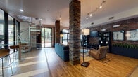 This club room is a well-appointed communal space that blends industrial character with modern comfort. Prominent exposed brick columns anchor the open floor plan, complementing warm hardwood flooring and track lighting overhead that creates a welcoming, well-lit atmosphere throughout.

The main lounge area is generously sized, furnished with a plush sectional sofa, leather accent chairs, and a wood-top coffee table arranged atop a woven area rug. Large glass doors open directly to an outdoor terrace, flooding the space with natural light and providing easy access to the exterior. A dedicated entertainment wall features a flat-screen television mounted above open shelving with decorative accents.

A wet bar and refreshment station offer added convenience, complete with a beverage refrigerator, microwave, marble-style countertops, and a hot beverage dispenser stocked with a variety of tea selections — ideal for casual gatherings or a quick break.

A dramatic spiral staircase serves as both a functional and sculptural focal point, adding vertical interest to the space. Adjacent to the lounge, a fitness center is visible through full-height glass walls and is outfitted with strength and cardio equipment, including cable machines, a bench press, treadmills, and stationary bikes, all beneath ceiling fans and bright overhead lighting.