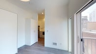 This single-floor space showcases a clean, contemporary aesthetic defined by crisp white walls, recessed lighting, and light-toned hardwood flooring that runs throughout. The layout is thoughtfully organized, with the main living area flowing seamlessly into the kitchen through an open passageway. Tall ceilings enhance the sense of openness, while a built-in white wardrobe or storage cabinet provides practical, integrated storage within the living space. A floor-level vent and wall-mounted thermostat indicate a dedicated climate control system for year-round comfort. Natural light filters in through a window on the right side of the living area, contributing to the bright and airy atmosphere. The kitchen, visible through the connecting opening, is outfitted with gray cabinetry, a stainless steel refrigerator, a built-in oven, and white upper cabinets — offering a functional and modern culinary space. The overall design emphasizes simplicity, efficiency, and a neutral color palette, making the space feel polished and move-in ready.