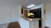 This single-story residence offers three bedrooms and two bathrooms within a well-organized floor plan. The kitchen features dark espresso-toned cabinetry that contrasts with light countertops, and is equipped with black appliances including a refrigerator and range. A peninsula-style counter extends into the adjacent area, providing additional prep space and a natural transition into the living and dining areas. The entryway is positioned directly off the kitchen, with a solid panel front door providing access to the unit. Flooring in the main living and entry areas consists of hardwood-style planking, while the living room is finished with carpet, adding warmth underfoot. The living room is a generously sized space featuring neutral white walls, a textured ceiling, and a window fitted with horizontal blinds that allows natural light to enter. A hallway connects the living areas to the bedrooms and bathrooms beyond, with overhead lighting illuminating the corridor. Throughout the space, a clean, neutral color palette creates a bright and functional environment well-suited for everyday living.