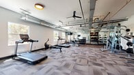 Located at 301 W Roosevelt St in Phoenix, Arizona, this fitness center offers a well-equipped, single-floor workout facility designed to accommodate a wide range of exercise routines. The space features an open, airy layout beneath an exposed industrial-style ceiling with visible ductwork, ceiling fans for air circulation, and warm overhead lighting that creates an energetic yet comfortable atmosphere.

The cardio section includes treadmills, a rowing machine, and an elliptical trainer, complemented by wall-mounted flat-screen TVs. Natural light filters in through multiple windows, with views of the surrounding Phoenix streetscape, including glimpses of palm trees that reinforce the Arizona setting.

The strength training area is comprehensively outfitted with a Smith machine, squat rack, adjustable bench, and an extensive dumbbell rack spanning a wide range of weights. A dedicated TRX suspension training station and cable machine add functional fitness options. Full-length mirrors line portions of the walls, enhancing the sense of space and supporting proper form during workouts.

Throughout the facility, durable patterned carpet tiles cover the floor, providing both comfort and traction. A storage shelving unit offers organized space for personal belongings. The overall design balances industrial aesthetics with practical functionality, making this fitness center a comprehensive resource for residents in the heart of downtown Phoenix.