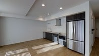 This single-floor space showcases a clean, contemporary design with an open-concept layout that seamlessly integrates the kitchen and living areas. The kitchen is appointed with a full suite of stainless steel appliances, including a French door refrigerator, built-in oven, microwave, dishwasher, and a cooktop with a stainless steel range hood above. Dark espresso-toned cabinetry provides a striking contrast against the crisp white subway tile backsplash that extends the full length of the kitchen wall, lending a modern yet timeless aesthetic. A mounted rail system above the countertop offers additional functional storage. The countertops complement the dark cabinetry for a cohesive look throughout the kitchen. Light hardwood flooring runs continuously across the space, enhancing the sense of openness and warmth. Recessed lighting in the ceiling provides even, ambient illumination throughout, while natural light streams in from windows, casting soft patterns across the floor. The walls are finished in a neutral white, keeping the interior bright and versatile. The overall ambiance is minimalist and functional, suited to a variety of uses.