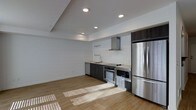 This single-floor space showcases a clean, contemporary design with an open-concept layout that seamlessly integrates the kitchen and living areas. The kitchen is appointed with a full suite of stainless steel appliances, including a French door refrigerator, built-in oven, microwave, dishwasher, and a cooktop with a stainless steel range hood above. Dark espresso-toned cabinetry provides a striking contrast against the crisp white subway tile backsplash that extends the full length of the kitchen wall, lending a modern yet timeless aesthetic. A mounted rail system above the countertop offers additional functional storage. The countertops complement the dark cabinetry for a cohesive look throughout the kitchen. Light hardwood flooring runs continuously across the space, enhancing the sense of openness and warmth. Recessed lighting in the ceiling provides even, ambient illumination throughout, while natural light streams in from windows, casting soft patterns across the floor. The walls are finished in a neutral white, keeping the interior bright and versatile. The overall ambiance is minimalist and functional, suited to a variety of uses.