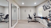 Located at 2096 E Main St in Spartanburg, South Carolina, this single-floor fitness facility is designed to provide a clean, functional, and motivating workout environment. The space is finished with light hardwood-style flooring throughout, neutral gray walls, and recessed ceiling lighting that keeps the area bright and welcoming.

The facility is equipped with a well-rounded selection of True-brand cardio machines, including treadmills, elliptical trainers, a stair climber, and both upright and recumbent stationary bikes. Multiple large, black-framed floor-to-ceiling mirrors are positioned along the walls to enhance the sense of space and allow users to monitor their form during exercise.

A standout design element is the bold motivational typography painted directly on the wall, reading "Will it be easy? Nope. Worth it? Absolutely." — a detail that adds character and energy to the workout environment. Natural light filters in through several windows fitted with white horizontal blinds, contributing to an open and airy atmosphere.

Additional wall-mounted accessories, such as equipment hooks, are present throughout, supporting versatility and organization. A set of white storage closets provides practical utility space. The overall design is modern, uncluttered, and purpose-built to support a focused and effective fitness experience.