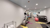The Studios at Patchen offers residents access to a dedicated fitness center designed to support a well-rounded exercise routine. The room is finished with clean white walls and a vibrant patterned carpet featuring bold yellow and pink accents, giving the space an energetic and modern aesthetic. Recessed lighting throughout the ceiling ensures the area is well-lit at all times, while a full-length mirrored wall on one side visually expands the space and allows for form-checking during workouts. The fitness center is equipped with a variety of cardio machines, including an elliptical trainer and treadmills, catering to a range of fitness preferences. A colorful collection of medicine balls is neatly arranged along one wall, alongside additional strength and conditioning equipment. Exercise stability balls are also available for core training and stretching. A metal utility shelf near the entrance provides convenient storage for towels and accessories. The layout is thoughtfully organized to allow for comfortable movement between equipment. The overall ambiance is clean, functional, and inviting, reflecting a commitment to resident wellness and an active lifestyle.