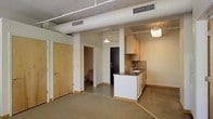 This single-floor space showcases a modern, open-concept layout that blends industrial and contemporary design elements. The exposed ceiling with visible ductwork and HVAC vents adds an urban loft aesthetic, while pendant lighting fixtures provide warm, focused illumination throughout.

The kitchen is positioned at the heart of the space, featuring a peninsular island with a dark countertop and integrated sink, light wood upper cabinetry, and a tile backsplash. The kitchen flows naturally into the adjacent living area, which is finished with carpet flooring, creating a comfortable and distinct zone within the open plan.

Polished concrete flooring in the kitchen and transitional areas provides a durable, clean contrast to the carpeted living space. Light wood doors and trim throughout offer a warm counterpoint to the otherwise neutral palette of white walls and grey ceiling.

A hallway visible in the background leads to additional areas of the unit, including what appears to be a bathroom, adding to the overall functionality of the space. The layout is efficient and well-organized, making excellent use of the available square footage while maintaining an airy, uncluttered atmosphere well-suited for modern living.