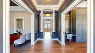 This thoughtfully designed single-floor space offers a refined blend of classic architectural detail and contemporary interior styling. The grand entrance hallway sets an impressive tone, featuring soaring ceilings, white classical columns framing a rotunda vestibule, rich hardwood flooring, and bold navy wainscoting accented with crisp white millwork. Pendant lighting with gold-toned fixtures adds warmth throughout the corridor, which leads visitors naturally into the heart of the space.

The living room serves as the focal centerpiece, showcasing a white marble fireplace surround flanked by built-in shelving and cabinetry. A dramatic botanical-patterned wallcovering in silver and gold provides a striking backdrop, while a circular halo pendant light and coffered ceiling detail elevate the room's sophistication. Large windows invite natural light, complementing the warm hardwood floors.

The combined dining and kitchen area includes a galley-style kitchen outfitted with white cabinetry, quartz countertops, stainless steel appliances, and a matte black faucet — functional yet polished in design.

The private office is well-appointed with natural light from multiple windows, decorative artwork, a conference-style round table, and dedicated desk space, making it ideal for focused work or consultations.

Connecting these areas is a welcoming hallway with curated artwork and glass-paned interior doors that maintain openness while defining each zone.