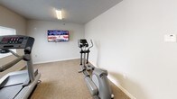 The fitness room is a single-level space designed to support basic cardiovascular exercise needs. The room features neutral-toned walls and durable, patterned carpet flooring throughout, providing a clean and functional environment. Two windows fitted with horizontal blinds allow natural light to filter into the space, contributing to an open and comfortable atmosphere during workouts.

The room is equipped with a treadmill and an elliptical machine, positioned to allow adequate clearance for safe use. A flat-screen television is mounted on the wall at an elevated viewing height, enabling users to watch programming while exercising. Overhead flush-mounted lighting provides consistent illumination throughout the space.

A wall-mounted HVAC unit ensures climate control within the room, maintaining a comfortable temperature year-round. Access is provided through a standard interior door with a windowed panel. The overall layout is straightforward and efficient, with open floor space that could accommodate additional equipment or stretching areas. The room's simple, no-frills design keeps the focus on functionality, making it a practical facility for everyday fitness use.