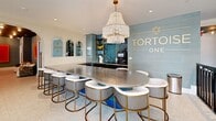 Located at 805 N Olive Ave in West Palm Beach, Florida, Tortoise One is a thoughtfully designed community space that seamlessly blends contemporary aesthetics with welcoming functionality. The single-floor layout encompasses a generous footprint organized into multiple distinct zones, each offering its own character and purpose.

At the heart of the space is a striking bar and lounge area anchored by a sweeping stainless steel countertop surrounded by white and gold upholstered barstools. Above, a tiered beaded chandelier draws the eye upward, while sea-glass teal grasscloth walls and brass-accented open shelving create a polished, coastal-modern backdrop. Arched mirror niches add architectural depth to the reception area.

Adjacent lounge zones feature warm reclaimed wood accent walls, a flat-screen television, plush sectional seating, and an eclectic mix of woven and upholstered chairs arranged around low tables. Bold, large-format artwork punctuates the blue-grey textured walls, adding vivid color and artistic energy throughout.

A dedicated conference or meeting room offers dark-topped tables, patterned tile flooring, and floor-to-ceiling windows branded with the Tortoise One logo, allowing ample natural light to filter in. The entry corridor is bright and open, lined with tall tropical plants and intimate café-style seating that creates a welcoming first impression. The overall ambiance is sophisticated yet approachable, reflecting a dynamic, design-forward community identity.