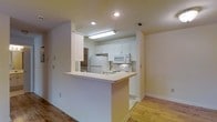 This single-story space showcases a well-designed open-concept kitchen that serves as a central feature. The kitchen is outfitted with crisp white cabinetry offering generous storage, complemented by white appliances including a refrigerator, electric range, and built-in microwave. A peninsula with a light-toned countertop extends into the living area, creating a functional breakfast bar that encourages casual dining and enhances the open flow between spaces. Recessed lighting and a flush-mounted ceiling fixture provide ample illumination throughout the kitchen area. Light hardwood-style flooring runs throughout, adding warmth and continuity to the space. The neutral color palette, featuring soft white walls and light-toned surfaces, creates a clean, bright atmosphere. The open layout between the kitchen and the adjoining living area promotes an airy, connected feel throughout. The space appears well-maintained and move-in ready, with practical features that balance everyday functionality with a tidy, welcoming aesthetic.
