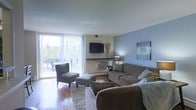 This single-floor space is designed with comfort and functionality in mind. The living room serves as the heart of the home, showcasing warm hardwood flooring that adds a classic, inviting character to the space. A soft, neutral area rug anchors the seating arrangement, which includes a generous sectional sofa and accent chairs, creating an ideal setting for relaxation and entertaining.

A calming blue accent wall adds a pop of color and visual interest, complemented by tasteful wall art. The wall-mounted flat-screen television serves as a focal point, positioned conveniently for viewing from multiple seating angles. A stylish floor lamp provides warm ambient lighting, enhancing the overall atmosphere of the room.

One of the most notable features of the living area is the large sliding glass door that floods the space with abundant natural light and offers direct access to an outdoor balcony. The balcony provides views of the surrounding outdoor environment, blending indoor and outdoor living seamlessly. Vertical blinds on the sliding door allow for adjustable privacy and light control.

Overall, the space presents a well-appointed, functional layout with a warm and welcoming ambiance, making it a comfortable and practical living environment.