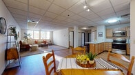 This single-floor unit offers a well-organized layout that balances open communal spaces with private rooms. The main living area is generously sized, featuring warm hardwood-style flooring, a drop tile ceiling with track lighting, and a bank of windows that frame views of the surrounding urban cityscape, filling the space with natural light. The living room transitions seamlessly into a defined dining area, which accommodates a round dining table and chairs on the same hardwood-style flooring.

The kitchen is positioned along one side of the open-plan area and features a peninsula counter with bar seating, wood-toned cabinetry, light countertops, a stainless steel refrigerator, and standard appliances including a microwave. Adjacent to the kitchen, a dedicated closet houses a stacked washer and dryer unit along with a water heater, offering convenient in-unit laundry.

The bedrooms are carpeted and quietly tucked away, each offering closet space and windows with urban outlooks. The primary bedroom is notably spacious with a walk-in closet area. The bathroom includes a tub-shower combination, a wood-toned vanity, and built-in shelving for storage. Throughout, the unit maintains a neutral, light palette.