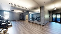 Located at 815 Court St in Utica, New York, this fitness facility offers a thoughtfully designed single-floor layout encompassing a generous amount of space. The interior blends modern finishes with industrial character, most notably expressed through exposed painted brick walls that lend texture and warmth throughout.

The primary exercise room is a well-appointed fitness space featuring light wood-tone luxury vinyl plank flooring, soft blue-gray painted walls, and bright overhead fluorescent lighting. It is outfitted with a comprehensive selection of cardio and strength equipment, including treadmills, a stationary bike, an elliptical, a cable machine, and a dumbbell rack, making it suitable for a full-body workout regimen.

A secondary open room serves as a flexible studio space, highlighted by floor-to-ceiling mirrors along one wall and exposed white-painted brick on the adjacent wall, creating a clean, open environment ideal for yoga, stretching, or group fitness activities. The same wood-tone flooring flows throughout, maintaining a cohesive aesthetic.

The hallway provides a polished entry experience, with cool gray walls, dark-trimmed glass doors, and access control panels that contribute to both security and a professional appearance. Natural light filters in through the glass entry doors and a window in the main exercise area, adding brightness to the overall atmosphere.