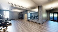 Located at 815 Court St in Utica, New York, this fitness facility offers a thoughtfully designed single-floor layout encompassing a generous amount of space. The interior blends modern finishes with industrial character, most notably expressed through exposed painted brick walls that lend texture and warmth throughout.

The primary exercise room is a well-appointed fitness space featuring light wood-tone luxury vinyl plank flooring, soft blue-gray painted walls, and bright overhead fluorescent lighting. It is outfitted with a comprehensive selection of cardio and strength equipment, including treadmills, a stationary bike, an elliptical, a cable machine, and a dumbbell rack, making it suitable for a full-body workout regimen.

A secondary open room serves as a flexible studio space, highlighted by floor-to-ceiling mirrors along one wall and exposed white-painted brick on the adjacent wall, creating a clean, open environment ideal for yoga, stretching, or group fitness activities. The same wood-tone flooring flows throughout, maintaining a cohesive aesthetic.

The hallway provides a polished entry experience, with cool gray walls, dark-trimmed glass doors, and access control panels that contribute to both security and a professional appearance. Natural light filters in through the glass entry doors and a window in the main exercise area, adding brightness to the overall atmosphere.