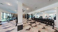 Located at 4126 River Rd in Wilmington, NC, this contemporary fitness center offers a thoughtfully designed, single-floor exercise space that provides a comprehensive workout environment. The layout is divided into distinct functional zones, each finished with a distinctive geometric-patterned carpet tile flooring that adds visual interest throughout the main training areas.

The cardio zone features a row of treadmills, a rowing machine, and a stair climber, all positioned along a windowed wall that floods the space with natural light and offers views to the outdoors. Ceiling fans provide additional comfort throughout the workout area.

The strength training section is generously equipped with a full dumbbell rack, multiple cable and resistance machines, and a variety of True-brand equipment. Floor-to-ceiling mirrors line the walls, enhancing the sense of openness and allowing users to monitor their form during exercises.

A dedicated studio area, separated from the main floor by a striking slatted wood partition wall, offers polished hardwood flooring, wall-to-wall mirrors, medicine balls, and space for stretching or group fitness activities. The warm wood tones of the partition provide an inviting contrast to the clean white walls and modern finishes found throughout. The overall ambiance is bright, clean, and professionally appointed, delivering a full-service fitness experience.