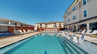 Located at 1226 Bexton St in Charlotte, NC, this outdoor amenity space centers around a generously sized resort-style swimming pool with crystal-clear blue water. The pool deck is thoughtfully designed to maximize relaxation and social enjoyment, lined on multiple sides with rows of modern white lounge chairs and sun loungers that invite residents to unwind under the Carolina sky.

Adjacent to the pool, a covered lounge area runs along the building's exterior, featuring large glass garage-style doors that open fully to blend indoor and outdoor living. String lights accent this transitional space, which is outfitted with additional lounge seating for shade and comfort.

A standout feature of the deck is a stylish wicker cabana daybed with a canopy and vibrant blue cushions, tucked against a classic brick accent wall — offering a private retreat within the communal space. Additional seating areas and colorful umbrellas are distributed throughout the deck, providing both sun and shade options.

The surrounding multi-story building features a contemporary design blending neutral siding, brick detailing, and dark metal awnings. With open sightlines, abundant natural light, and a well-appointed deck layout, this outdoor space delivers a compelling resort atmosphere in a residential setting in Charlotte's southwest corridor.