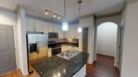 Located at 5201 Collin-McKinney Pkwy in McKinney, TX, this single-floor residence offers a thoughtfully designed layout with a blend of modern finishes and functional living spaces. The kitchen is a standout feature, showcasing rich dark granite countertops, white shaker-style cabinetry with brushed nickel hardware, and a full suite of stainless steel appliances including a French door refrigerator, electric range, and built-in microwave. A center island with a double-basin sink and elegant pendant lighting serves as a natural focal point, complemented by a stone tile backsplash and track lighting overhead. Warm hardwood flooring flows through the kitchen and living areas, accented by crown molding and arched doorway details that add architectural character. The bedrooms are carpeted and generously sized, each featuring ceiling fans and large windows that invite natural light and outdoor views. The primary bathroom is outfitted with an L-shaped double vanity topped with stone counters, tile flooring, and ample cabinetry. A secondary bathroom includes a full tub-shower combination with classic subway tile surround. The overall ambiance is clean, neutral, and move-in ready, reflecting a well-maintained and cohesively designed living environment.
