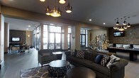 This stylish single-floor space exudes a modern, upscale ambiance with a carefully curated interior design. The lounge area is anchored by a generously sized dark tufted sofa adorned with patterned throw pillows, complemented by a matching accent chair and a sleek round coffee table resting atop a bold geometric-patterned area rug. A striking cluster of copper and bronze pendant lights hangs overhead, adding warmth and visual drama to the space. Recessed lighting further enhances the overall illumination throughout.

Large floor-to-ceiling windows and glass doors line the perimeter, inviting abundant natural light and offering clear views to an outdoor seating area beyond, creating a seamless indoor-outdoor connection. The neutral tones of the walls and ceiling are offset by rich, dark furnishings and eye-catching decorative accents, including a large sculptural sunburst piece in metallic gold that serves as a prominent focal point. The overall design palette blends deep grays, gold, and navy, lending the space a refined yet welcoming character. The open layout and thoughtful arrangement of seating make this an ideal setting for gathering, relaxing, or socializing in an elevated environment.