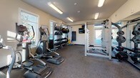 This fitness center is a comprehensively equipped single-floor facility designed to support a wide range of workout styles and fitness goals. The space features durable rubber flooring throughout, providing both safety and practicality for high-intensity exercise. Neutral-toned walls and flush fluorescent lighting create a clean, functional atmosphere, while multiple windows fitted with blinds allow natural light to filter in and offer views of the surrounding area.

The cardio zone is anchored by a row of Star Trac treadmills and elliptical machines, offering users reliable options for aerobic training. A dedicated strength area includes a multi-function cable machine and a power rack with weight plate storage, catering to both machine-based and free-weight training. Along one wall, a well-organized storage rack holds an assortment of dumbbells, medicine balls, TRX suspension trainers, resistance bands, and exercise mats, supporting functional and bodyweight-based workouts. A wall-mounted television provides entertainment or instructional content. A water cooler and posted workout guides add further convenience. Full-length mirrors enhance the sense of space while allowing users to monitor their form during exercise. The overall layout is open and thoughtfully arranged for efficient use of the available space.