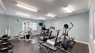Located at 100 Lochaven Drive in Charleston, South Carolina, this single-floor community amenity facility offers residents two functional spaces designed for everyday convenience and wellness. The exercise room is the centerpiece of the facility, finished with soft blue-gray walls, crown molding, and bright overhead fluorescent lighting that ensures a well-lit workout environment. Rubber flooring provides a durable, safety-conscious surface underfoot. The room is equipped with a multi-station cable weight machine, treadmills, an elliptical trainer, a stationary bike, and a free-weight rack, catering to a range of fitness routines. Full-length mirrors along one wall enhance the sense of space and allow for form monitoring during exercise. A wall-mounted television adds an entertainment option during workouts, and windows with blinds allow for natural light while maintaining privacy. Adjacent to the fitness area, the laundry room is outfitted with multiple commercial-grade washers and dryers lined along the walls, all set against the same cohesive blue-gray color palette and light wood-look flooring. A round dining table with chairs provides a comfortable waiting area, and a wall-mounted television keeps residents entertained. Informational signage is posted throughout for user guidance. Together, these spaces reflect a practical and well-maintained community amenity hub.