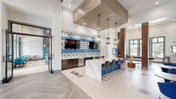 Located at 12810 Cypress Cape Cir in Fort Myers, Florida, this single-story community clubhouse offers a generously sized, open-concept layout designed for socializing and relaxation. The space is anchored by a striking main living area with soaring ceilings, large floor-to-ceiling windows that flood the interior with natural light, and views of the surrounding outdoor amenities including a pool. A herringbone-patterned wood-look floor runs through the central hub, lending a sophisticated yet welcoming character to the space.

The entrance lobby makes an immediate impression with vibrant tropical-themed artwork displayed against a deep navy accent wall, complemented by royal blue lounge seating and bistro-style tables. Bold, textured stone columns frame the transition between zones, adding an earthy warmth that balances the crisp white walls and modern furnishings.

A dedicated game room features a full-size pool table and a Jenga tower, while a wet bar and built-in cabinetry with a wall-mounted TV provide entertainment amenities nearby. The lounge area offers a variety of seating configurations — from teal sofas and accent chairs to intimate café tables — all bathed in ample natural light. A geometric patterned tile accent wall and pendant lighting fixtures add finishing touches that elevate the overall design. This clubhouse is a well-appointed, amenity-rich gathering space suited for community engagement.