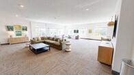 Located at 5102 Station Dr in Birmingham, Alabama, this generously proportioned single-floor living room serves as a well-appointed communal gathering space. The open-plan layout is bright and airy, thanks to an extensive array of large windows lining multiple walls, which flood the interior with natural light and offer pleasant views of the surrounding greenery.

The space is organized into several distinct functional zones. A primary lounge area anchors the room with a large, plush sectional sofa in warm olive tones, paired with a dark coffee table and a sculptural accent stool. Adjacent to this is a more intimate seating nook furnished with curved lavender chairs and a compact white table, ideal for casual conversation. A dedicated dining area features a round white table surrounded by woven rattan chairs beneath a warm pendant light, adding a welcoming, organic texture to the space.

An entertainment wall serves as a focal point, featuring dual wall-mounted flat-screen televisions set against a sophisticated grey accent wall, with a streamlined sectional sofa in front. Additional wall-mounted screens and a wood-paneled credenza provide further media functionality throughout the room. Patterned, textured carpeting runs throughout, complementing the neutral white walls and clean architectural lines. Colorful wall art adds personality and visual interest, completing this modern, versatile community space.