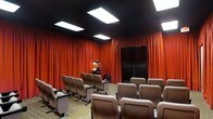 This private screening room delivers an authentic cinematic experience within a single, thoughtfully designed space. The room is anchored by rows of cushioned theater-style seats arranged in a tiered layout, ensuring unobstructed sightlines to the large flat-screen display mounted at the front. Deep crimson drapes line the walls from floor to ceiling, evoking the timeless ambiance of a classic movie house while also serving excellent acoustic purposes. The dark ceiling, fitted with recessed rectangular light panels, creates a dramatic overhead contrast and allows for precise lighting control to set the perfect viewing atmosphere. A popcorn machine stationed near the entrance adds a charming, authentic touch to the cinematic experience. The patterned carpet underfoot contributes to the room's sound dampening qualities while reinforcing the traditional theater aesthetic. Every element of this space has been carefully curated to immerse viewers in a dedicated entertainment environment, making it ideal for private film screenings, presentations, or group viewings. The combination of professional-grade seating, controlled lighting, and rich décor results in an intimate yet impressive venue that prioritizes both comfort and visual impact.