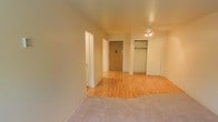 This single-floor space offers a straightforward and functional layout designed to maximize usability. The main living area features warm hardwood flooring that transitions seamlessly into a carpeted zone, creating a natural distinction between the living and sleeping areas. A ceiling fan with an integrated light fixture is centrally mounted overhead, providing both airflow and ambient lighting throughout the space. A wall-mounted air conditioning unit beneath a window ensures climate control, while the window itself allows natural light to filter into the room, brightening the interior. The neutral, light-toned walls contribute to an open and airy atmosphere throughout. Toward the entry area, a solid front door is flanked by a built-in closet outfitted with a hanging rod and shelving, offering practical storage directly accessible from the main living space. An additional doorway leads to a separate room, expanding the overall functionality of the unit. The clean lines, minimal architectural detailing, and combination of flooring materials give the space a simple yet versatile character, well-suited for a variety of furnishing arrangements and living needs.