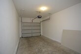 This single-level garage presents a clean, functional layout well-suited for vehicle storage, utility use, or workshop activities. The space is finished with smooth white walls that lend a bright, tidy appearance throughout. The concrete floor is durable and easy to maintain, providing a solid foundation across the full extent of the garage. A motorized overhead door opener is centrally mounted to the ceiling, paired with a sectional roll-up garage door that features metal panel detailing and offers convenient vehicle access. The ceiling is finished with a textured surface and includes integrated lighting that evenly illuminates the interior. An interior door along the back wall provides direct access to the adjoining property, enhancing everyday convenience. The overhead door track system runs along the ceiling, and the overall layout is open and unobstructed, maximizing usable floor space. The neutral color palette and clean lines give the space a well-kept, move-in-ready feel. Whether used for parking, additional storage, or a hobby workspace, this garage offers a versatile and practical environment.