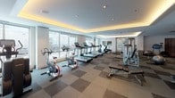 This fitness center offers a thoughtfully designed, open-plan workout space spread across a single level. The room is finished with a distinctive checkered flooring pattern in neutral tones, providing a clean and organized visual foundation throughout. Elegant cove lighting runs along the perimeter of the ceiling, casting a warm, ambient glow that complements the recessed pot lights and contributes to an inviting atmosphere.

Large windows along one wall face the street, flooding the cardio zone with natural light and providing views of the surrounding area. This section is equipped with multiple treadmills, recumbent and upright stationary bikes, and cycling bikes, all positioned to take advantage of the natural light and outdoor views.

The strength training area occupies a generous portion of the floor plan and includes a variety of cable and resistance machines, a dumbbell rack, adjustable benches, and stability balls. A wall-mounted flat-screen television is positioned to be visible from multiple workout stations.

Additional amenities include a water fountain station and towel service area. White walls, smooth ceilings, and a modern, neutral color palette throughout give the space a polished, contemporary feel well-suited to a full workout routine.