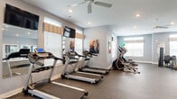 Located at 3820 Codet Rd in Columbus, Ohio, this single-level fitness center offers a comprehensive and thoughtfully organized workout environment. The space is divided into distinct functional zones, providing dedicated areas for cardio training, strength work, and open-floor exercise.

The primary cardio area is generously proportioned, housing multiple treadmills and elliptical machines arranged in orderly rows. Wall-mounted flat-screen televisions face the treadmills, and large mirrors along the walls enhance the sense of space while allowing users to monitor their form. Multiple windows fitted with decorative valances and blinds admit ample natural light throughout this zone, while ceiling fans contribute to air circulation and comfort. Recessed lighting supplements the natural light for consistent illumination.

A separate open exercise area provides ample floor space for stretching, functional training, or group workouts. This room features the same durable rubber-composite flooring found throughout the facility, soft gray walls, and additional ceiling fan coverage. Strength equipment and a cable machine are positioned here, alongside a rack of stability balls in varying sizes. Interior windows and glass panels connect this space visually to the adjacent rooms.

Motivational fitness artwork displayed on the walls adds an energetic ambiance, reinforcing the facility's active, wellness-focused character throughout.
