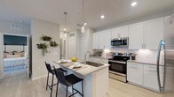 This well-designed single-floor residence showcases a contemporary aesthetic throughout, with light wood-style flooring, neutral tones, and recessed lighting creating a cohesive and inviting atmosphere. The kitchen is a standout feature, outfitted with crisp white shaker cabinetry, a white subway tile backsplash, and a full suite of stainless steel appliances including a refrigerator, range, and built-in microwave. A central island with a dual-basin sink and quartz-style countertop provides casual seating for two, illuminated by sleek pendant lights overhead. The open living area offers generous natural light through multiple windows dressed with sheer white curtains and blinds. A ceiling fan adds comfort, while light flooring and neutral walls create a versatile, airy backdrop. The primary bedroom features plush carpet, a bold accent wall, ceiling fan, natural light from a window with blinds, and direct access to a spacious walk-in closet. A secondary bedroom echoes the same comfortable carpet flooring and neutral palette. The bathroom is finished with white cabinetry, a grey countertop vanity, a large mirror, and a full tub-shower combination, maintaining the clean, modern style found throughout the home.
