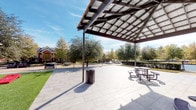 The Grilling Pavilion is a well-appointed outdoor communal space designed for gatherings, leisure, and al fresco dining. At its centerpiece is a generously sized covered pavilion with a striking modern overhead structure composed of dark metal framing and a grid-patterned roof that provides shade while maintaining an open, airy atmosphere. The patio beneath is finished with large-format light gray tiles, offering a clean and durable surface that comfortably accommodates multiple picnic tables and seating areas. A decorative perforated metal waste receptacle is conveniently positioned within the space. Surrounding the pavilion, patches of manicured turf and gravel landscaping provide visual contrast and low-maintenance greenery. Mature trees frame the area, offering natural shade and seasonal color, while views of the broader residential community are visible in the background. Adjacent to the pavilion, a building with classic brick and lap siding exterior is visible, along with a garage door and concrete walkway, suggesting convenient access from nearby residences. The overall setting is open, bright, and welcoming, making it a practical and attractive amenity for outdoor cooking, socializing, and community engagement throughout the seasons.