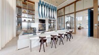 This elegant single-floor amenity space is designed with a refined, contemporary aesthetic that seamlessly blends form and function. Soaring double-height ceilings create an expansive, airy atmosphere, while floor-to-ceiling steel-framed windows flood the interior with abundant natural light and offer sweeping views of a resort-style pool and surrounding palm-lined grounds.

The social lounge area is anchored by a striking fireplace set against a textured accent wall, above which a wall-mounted display screen is integrated into the design. A long, polished marble-top bar counter runs parallel to the fireplace, attended by sculptural dark wood bar stools, making it an ideal gathering spot. A dramatic sculptural chandelier with fluid blue pendants hangs overhead, serving as a bold focal point.

Built-in wood shelving units flank the feature wall, displaying curated décor and providing generous storage. A fully equipped kitchenette area includes a double wall oven, stainless steel dishwasher, refrigerator, a sink with quartz countertops, and a wall-mounted television set against a dimensional wave-textured tile backsplash.

Light-toned wide-plank hardwood flooring runs throughout, complementing the warm wood tones of the cabinetry and shelving. A large dining table with upholstered seating rounds out the space, providing a versatile area for gatherings and events. The overall ambiance is one of upscale sophistication and welcoming comfort.
