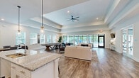 Located at 4605 Sidbury Crossing Dr in Castle Hayne, NC, this single-floor community clubhouse offers a thoughtfully designed open-concept layout that seamlessly blends a fully equipped kitchen with a spacious living and recreational area. The kitchen features a large granite-topped island with bar seating, white shaker cabinetry, stainless steel appliances, and elegant pendant lighting — ideal for gatherings and events. Wide-plank wood-look flooring flows throughout, complementing the bright, neutral color palette and clean architectural lines. A coffered ceiling with a ceiling fan anchors the living area, which is furnished with comfortable seating and a full-size pool table, creating a versatile social hub. Floor-to-ceiling black-framed glass doors and windows flood the space with natural light and open directly to an outdoor pool area surrounded by lush greenery. Additional areas include a dedicated dining space, a glass-enclosed fitness or office room, and a welcoming entryway with coastal-inspired wall décor. Wall-mounted televisions and recessed lighting are featured throughout. The overall ambiance is fresh, airy, and community-oriented, reflecting a contemporary coastal aesthetic well-suited to the North Carolina setting.