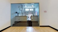 This fitness facility is defined by its striking glass-walled entrance, where frameless glass panels create a seamless, open visual connection between the exterior corridor and the gym interior. The space is accessed through glass doors that open to a split-level layout, with a short staircase leading up to the main workout area. The lower entry level features light hardwood flooring, while the elevated gym floor is finished in a darker material, providing a clear visual distinction between zones. Inside, a variety of exercise equipment is visible, including cardio machines and strength training apparatus. The interior walls are painted in neutral tones, complementing the clean, contemporary design. Elevated clerestory windows line the upper portion of the walls, flooding the space with natural light while maintaining privacy. Flush ceiling light panels provide additional illumination throughout. A mounted flat-screen display and framed artwork add functional and aesthetic touches to the walls. The double-height ceiling in the gym area enhances the sense of openness and volume, contributing to an airy, professional atmosphere well-suited for an active fitness environment.