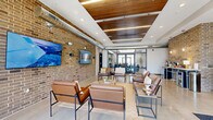 Located at 2290 SE Waco Pl in Waukee, Iowa, this generously sized single-floor amenity space blends industrial-modern design with warm, functional interiors. Exposed brick walls, polished concrete floors, and visible ductwork create a contemporary industrial aesthetic throughout, while wood-accented ceilings and soft lighting add warmth and character.

The main living and lounge area serves as the heart of the space, featuring comfortable leather seating arrangements, large-format mounted televisions, and a snack and beverage station. Natural light pours in through expansive windows and glass-panel doors that frame outdoor views of the surrounding Iowa landscape. A secondary living room offers additional seating with a cozy, relaxed feel, anchored by another mounted display set against an exposed brick accent wall.

The kitchen area is thoughtfully appointed with rich dark wood cabinetry, white countertops, stainless steel appliances, and a peninsula bar with industrial-style metal barstools. Pendant lighting over the bar and a wood plank ceiling lend a refined yet casual atmosphere.

The dedicated exercise room is well-equipped with cardio machines, free weights, adjustable benches, and a ceiling fan, all set on durable rubber flooring with ample natural light from large windows.

The hallway features built-in package lockers along both walls, offering a practical and organized solution for deliveries, completing this well-rounded communal facility.