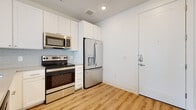 This single-floor unit showcases a clean, contemporary design with light hardwood-style flooring and crisp white walls carried consistently throughout the space. The floor plan, visible in the 3D layout, reveals a well-organized arrangement of living, kitchen, bedroom, and bathroom areas.

The kitchen is a standout feature, equipped with a full suite of stainless steel appliances, including a French door refrigerator with water and ice dispenser, an electric range, and an over-the-range microwave. White shaker-style cabinetry extends to the ceiling, providing generous storage, while a light blue tile backsplash adds a subtle decorative touch. The countertops offer ample prep space, and a single-basin sink with a modern faucet completes the workspace.

The bathroom features a tub-shower combination with white subway tile surround, brushed nickel fixtures, and a white vanity with a neutral-toned countertop, mirror, and additional upper cabinetry for storage. The bedroom is bright and airy, with a large window fitted with horizontal blinds allowing natural light, a ceiling fan, and a closet with built-in shelving nearby. The entry door opens directly adjacent to the kitchen, creating a compact yet functional layout.