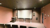 This single-floor screening room is purpose-built for an immersive cinematic experience. At the front of the room, a large, centrally mounted projection screen dominates the wall, framed cleanly against a dark brown backdrop. A stanchion barrier separates the screen area from the seating section, maintaining a structured viewing environment. The room is equipped with a full surround sound system, with wall-mounted speakers positioned on both sides of the screen as well as along the side walls, ensuring balanced audio throughout the space. A substantial subwoofer unit is placed to the left of the screen, contributing to a rich, full-bodied sound experience. Recessed lighting is evenly distributed across the dark drop ceiling, providing controlled ambient illumination that enhances the theatrical atmosphere. The flooring is carpeted in a neutral tone, and the seating consists of individual black chairs arranged in rows for optimal viewing angles. The overall design is intentional and functional, with dark tones on the ceiling and walls minimizing light reflection and keeping focus on the screen. This space is well-suited for private film screenings, presentations, or media events.