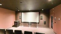 This single-floor screening room is purpose-built for an immersive cinematic experience. At the front of the room, a large, centrally mounted projection screen dominates the wall, framed cleanly against a dark brown backdrop. A stanchion barrier separates the screen area from the seating section, maintaining a structured viewing environment. The room is equipped with a full surround sound system, with wall-mounted speakers positioned on both sides of the screen as well as along the side walls, ensuring balanced audio throughout the space. A substantial subwoofer unit is placed to the left of the screen, contributing to a rich, full-bodied sound experience. Recessed lighting is evenly distributed across the dark drop ceiling, providing controlled ambient illumination that enhances the theatrical atmosphere. The flooring is carpeted in a neutral tone, and the seating consists of individual black chairs arranged in rows for optimal viewing angles. The overall design is intentional and functional, with dark tones on the ceiling and walls minimizing light reflection and keeping focus on the screen. This space is well-suited for private film screenings, presentations, or media events.