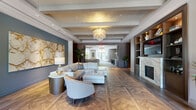 Located at 40 Pinehills Drive in Plymouth, Massachusetts, this refined single-floor space offers a generously proportioned layout defined by high-end architectural details and contemporary design throughout. The expansive living room serves as the centerpiece, showcasing a striking herringbone and parquet-patterned hardwood floor that anchors the entire space with warmth and texture. A coffered ceiling with a metallic gold finish adds architectural depth overhead, while a cascading capiz shell chandelier provides a sculptural focal point and soft, ambient illumination. A large-format marble artwork adorns a bold slate-blue accent wall, lending an artistic and sophisticated character to the room. Built-in shelving with warm wood tones flanks a marble-surround fireplace, offering both function and refined aesthetic appeal. Plush neutral sectional seating and elegant accent chairs create a welcoming and comfortable gathering area. The space flows seamlessly into a dedicated dining and kitchen area, featuring a substantial island with bar seating, pendant lighting, and high-end appliances—ideal for entertaining. The connecting hallway is finished with wide-plank hardwood flooring and leads to a glass-fronted entry, allowing natural light to filter through the space. Tall ceilings, recessed lighting, and lush potted greenery throughout contribute to an overall ambiance that is both polished and inviting.