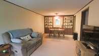 This single-floor home offers a welcoming and functional layout designed for comfortable everyday living. The combined living and dining area serves as the heart of the space, creating an open and cohesive environment that encourages both relaxation and gathering. The living area features plush wall-to-wall carpeting in a neutral tone, providing a soft and inviting atmosphere underfoot. A comfortable reclining sofa anchors the seating space, complemented by warm, ambient lighting overhead. The dining area is defined by a classic wooden table and chair set, positioned beneath a ceiling-mounted light fixture that adds a soft glow to the room. A standout feature of this space is the expansive built-in shelving unit that lines the wall between the living and dining areas. Finished in a rich, dark wood tone, the shelving provides generous display and storage space, adding both character and practicality to the interior. A window situated at the center of the shelving unit allows natural light to filter into the dining area, brightening the space. Crown molding along the ceiling adds a subtle architectural detail that lends a polished, finished look to the interior. Overall, the space presents a cozy and well-organized home with classic design sensibilities.