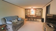 This single-floor home offers a welcoming and functional layout designed for comfortable everyday living. The combined living and dining area serves as the heart of the space, creating an open and cohesive environment that encourages both relaxation and gathering. The living area features plush wall-to-wall carpeting in a neutral tone, providing a soft and inviting atmosphere underfoot. A comfortable reclining sofa anchors the seating space, complemented by warm, ambient lighting overhead. The dining area is defined by a classic wooden table and chair set, positioned beneath a ceiling-mounted light fixture that adds a soft glow to the room. A standout feature of this space is the expansive built-in shelving unit that lines the wall between the living and dining areas. Finished in a rich, dark wood tone, the shelving provides generous display and storage space, adding both character and practicality to the interior. A window situated at the center of the shelving unit allows natural light to filter into the dining area, brightening the space. Crown molding along the ceiling adds a subtle architectural detail that lends a polished, finished look to the interior. Overall, the space presents a cozy and well-organized home with classic design sensibilities.