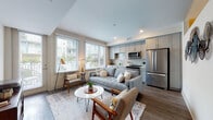 Located at 10 Westedge Street in Charleston, South Carolina, this single-floor residence offers a thoughtfully designed layout with contemporary finishes throughout. The open-concept living and kitchen area serves as the heart of the home, filled with natural light from a bank of large windows that offer views of the surrounding neighborhood. Rich hardwood-style flooring runs throughout the main living spaces, complementing the neutral palette of the walls and trim.

The kitchen is well-appointed with grey shaker-style cabinetry extending to the ceiling, a classic subway tile backsplash, light countertops, and a full suite of stainless steel appliances including a French door refrigerator, electric range with microwave above, and dishwasher. The living area provides ample room for comfortable seating arrangements, with recessed lighting adding warmth to the space.

A clean, well-lit hallway connects the living area to the private quarters. The bedroom features hardwood-style flooring, a reach-in closet with built-in shelving, and a ceiling fan. Conveniently located nearby is a full bathroom with tile flooring, a combination tub and shower, and a vanity with grey cabinetry. A stacked washer and dryer unit rounds out the in-unit amenities, offering exceptional everyday convenience.