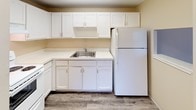 Located at 6935 York Rd in Parma Heights, Ohio, this single-floor unit offers a practical and functional layout designed for comfortable everyday living. The floor plan, as revealed by the 3D model, flows through several well-defined spaces connected in a compact, efficient arrangement.

The living area is carpeted in neutral gray tones and features warm beige walls, a ceiling fan with light fixture, and a pass-through window that opens directly to the kitchen — a design detail that promotes an open, connected feel between the two spaces. Natural light filters in through windows fitted with vertical blinds.

The kitchen is equipped with white cabinetry offering generous storage above and below the countertops, light-colored laminate counters, a stainless steel sink, and a full-size refrigerator. The flooring here transitions to wood-look vinyl planking, providing easy maintenance.

The bedroom is a quiet, carpeted retreat with warm beige walls, a window with views of the surrounding greenery, and a baseboard heating system. A wall-mounted air conditioning unit ensures year-round comfort.

The bathroom is clean and bright, finished in white tile surrounding a full soaking tub and shower combination, complemented by chrome fixtures. The overall ambiance throughout the unit is neutral, tidy, and move-in ready, making it a straightforward and functional living space.