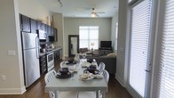 This single-floor residence features an open-concept layout that seamlessly connects the kitchen, dining, and living areas into one cohesive living space. The kitchen is outfitted with dark cabinetry, granite countertops, stainless steel appliances including a range and built-in microwave, and under-cabinet lighting, offering both functionality and a sleek aesthetic. Track lighting overhead adds to the modern feel.

The dining area sits centrally within the open floor plan and accommodates a white rectangular table with matching white chairs, providing a clean contrast to the dark hardwood flooring that runs throughout the space. The living area beyond features a comfortable sectional sofa, flat-screen television, and a warm lamp, creating a relaxed atmosphere. A ceiling fan with a light fixture serves the combined living and dining space efficiently.

Large windows dressed with white blinds line the space, allowing generous natural light to fill the interior. A floor-length mirror near the kitchen enhances the sense of openness. The overall ambiance is modern and practical, with a neutral color palette anchored by rich dark floors.