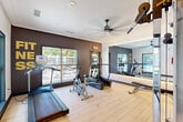 Located at 3400 Peachtree Corners Circle in Peachtree Corners, GA, this fitness center is a well-appointed exercise room designed to support a complete workout experience. The space features light hardwood-style flooring throughout, crown molding, and a clean white ceiling with recessed lighting and a modern ceiling fan to maintain airflow during workouts. A standout design element is the large backlit wall mirror, which visually expands the room and enhances the bright, energetic atmosphere. The accent wall, finished in a deep charcoal textured wallcovering, is adorned with bold gold "FITNESS" lettering, adding a motivational and stylish focal point. Cardio equipment includes a treadmill, elliptical, and stationary bike, while strength training is supported by a multi-use cable machine and an adjustable weight bench. Multiple windows along the exterior wall flood the space with natural light and offer views of a pool and outdoor recreation area, creating a refreshing connection to the outdoors. A glazed entry door provides easy access and visibility into the room. The overall ambiance is modern, clean, and functional, making it a practical and inviting space for daily exercise.